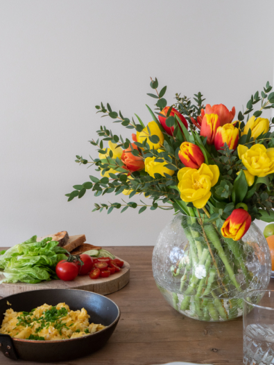 Kuvassa ruokapöydällä on iso tulppaanikimppu, jossa on keltasen, oranssin ja ounaisen sävyisiä tulppaaneja.