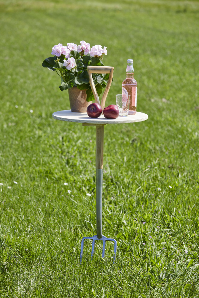 Kuvassa on valmis pöytä nurmikolla. Pöydällä on pelargoni ja pullo limonadia.